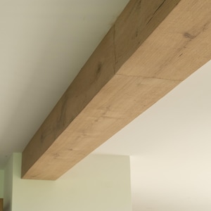 Puede incluir: Una gran viga de madera rectangular se extiende diagonalmente por la imagen. La viga tiene vetas de madera natural y un color marr&oacute;n claro. El fondo es un techo blanco y una pared verde claro.