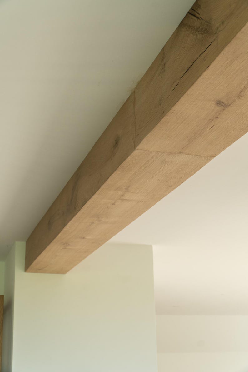 Puede incluir: Una gran viga de madera rectangular se extiende diagonalmente por la imagen. La viga tiene vetas de madera natural y un color marr&oacute;n claro. El fondo es un techo blanco y una pared verde claro.