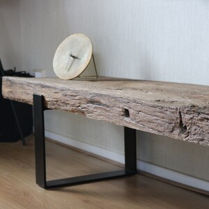 Puede incluir: Un banco largo con un asiento de losa de madera marr&oacute;n de bordes naturales y textura rugosa y desgastada. El asiento est&aacute; sostenido por dos patas de metal negro en forma de U. Un reloj circular con esfera clara se encuentra encima.