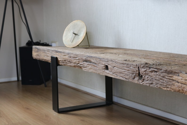 Puede incluir: Un banco largo con un asiento de losa de madera marr&oacute;n de bordes naturales y textura rugosa y desgastada. El asiento est&aacute; sostenido por dos patas de metal negro en forma de U. Un reloj circular con esfera clara se encuentra encima.