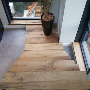 Op de afbeelding: Een houten trap met een lichtbruine afwerking. De treden zijn gemaakt van brede planken en hebben een natuurlijke, rustieke uitstraling. Een plant in een pot staat op een bordes boven de trap.