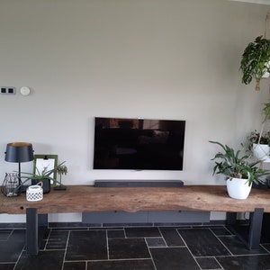 Puede incluir: Una mesa consola de madera larga y rectangular con un borde natural y dos patas de metal oscuro en forma de U. La mesa est&aacute; situada debajo de un televisor montado en la pared y una barra de sonido, con varios objetos decorativos y plantas.
