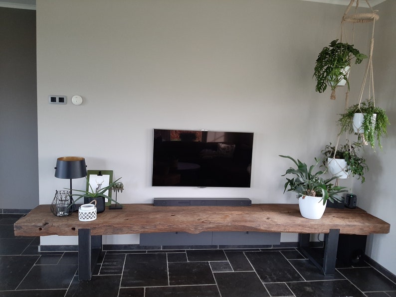 Puede incluir: Una mesa consola de madera larga y rectangular con un borde natural y dos patas de metal oscuro en forma de U. La mesa est&aacute; situada debajo de un televisor montado en la pared y una barra de sonido, con varios objetos decorativos y plantas.