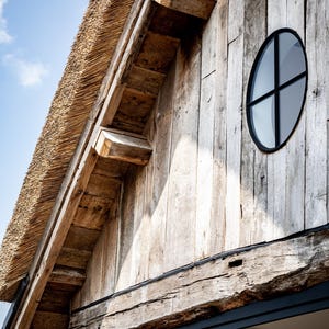 May include: Exterior view of a rustic building with a thatched roof and weathered wooden siding. A round window with a black frame is visible. The building's design incorporates natural materials and a classic aesthetic.