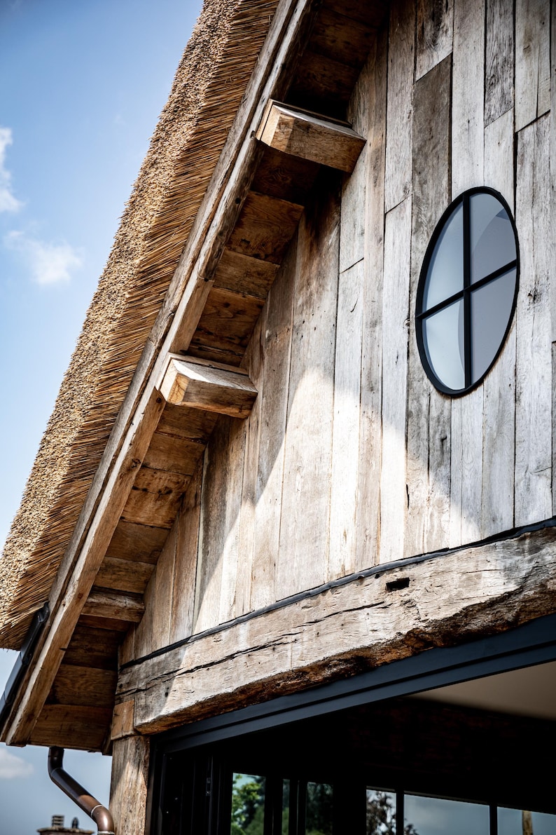 May include: Exterior view of a rustic building with a thatched roof and weathered wooden siding. A round window with a black frame is visible. The building's design incorporates natural materials and a classic aesthetic.