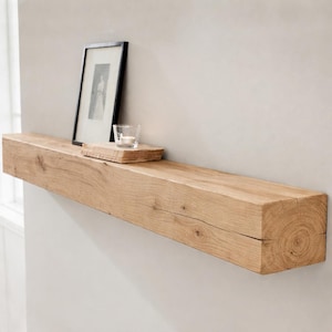Puede incluir: Una estantería flotante de madera natural, montada en una pared gris claro. Una foto en blanco y negro enmarcada, un pequeño bloque de madera y un portavelas de cristal con una vela encendida están sobre la estantería. La estantería mide unos 152 cm de largo.