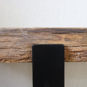 Puede incluir: Una pieza horizontal de madera clara desgastada con una veta rugosa y texturizada con grietas visibles, asegurada por un soporte de metal negro s&oacute;lido y liso que se extiende verticalmente desde el borde inferior de la viga de madera.