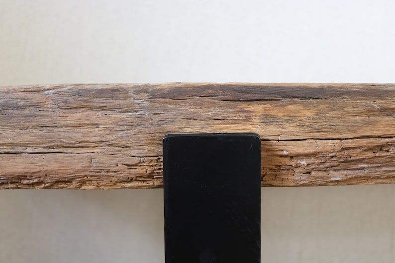 Puede incluir: Una pieza horizontal de madera clara desgastada con una veta rugosa y texturizada con grietas visibles, asegurada por un soporte de metal negro s&oacute;lido y liso que se extiende verticalmente desde el borde inferior de la viga de madera.