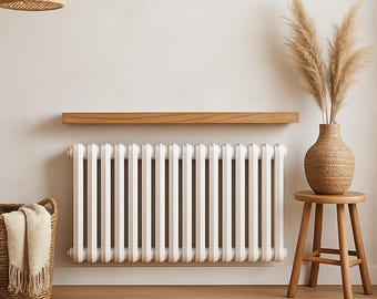 May include: A white, vertical radiator dominates the scene. A wooden shelf is mounted above it. A woven basket with a blanket, a round rug, a vase with dried plants, and a small wooden stool complete the neutral-toned, minimalist decor.