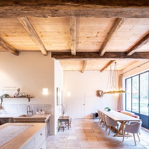 May include: A spacious kitchen and dining area with a rustic aesthetic. The room features a wooden ceiling with exposed beams, a large window, and a long wooden dining table. The kitchen has wooden cabinets and a large island.