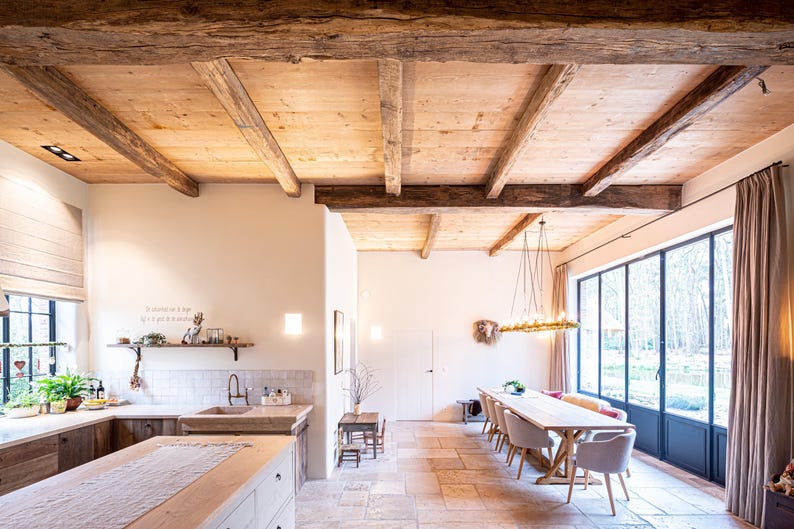 May include: A spacious kitchen and dining area with a rustic aesthetic. The room features a wooden ceiling with exposed beams, a large window, and a long wooden dining table. The kitchen has wooden cabinets and a large island.