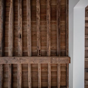 May include: A close-up view of a wooden ceiling with exposed beams. The ceiling is constructed of vertical and horizontal wooden beams, creating a grid-like pattern. The wood has a natural, aged appearance with varying shades of brown.