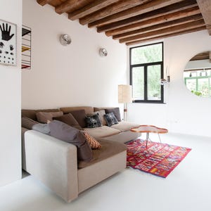 May include: A modern living room with a beige sectional sofa, decorative pillows, and a colorful rug. The room features a wooden beam ceiling, a round mirror, and a small wooden table. The walls are white, and the floor is also white.
