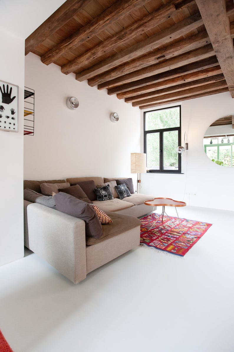 May include: A modern living room with a beige sectional sofa, decorative pillows, and a colorful rug. The room features a wooden beam ceiling, a round mirror, and a small wooden table. The walls are white, and the floor is also white.