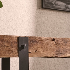 Puede incluir: Una mesa consola rectangular con un tablero de viga de madera marr&oacute;n desgastada con vetas y grietas visibles. La viga est&aacute; sostenida por dos soportes verticales de metal negro asegurados con pernos circulares prominentes en la parte frontal.