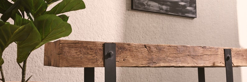 Puede incluir: Una mesa consola rectangular con un tablero de viga de madera marr&oacute;n desgastada con vetas y grietas visibles. La viga est&aacute; sostenida por dos soportes verticales de metal negro asegurados con pernos circulares prominentes en la parte frontal.