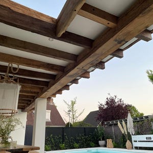 May include: An outdoor patio with a swimming pool. The patio features a wooden pergola with exposed beams and white panels. A wooden table and bench are in the foreground. The pool has clear blue water, and the background includes houses and greenery.