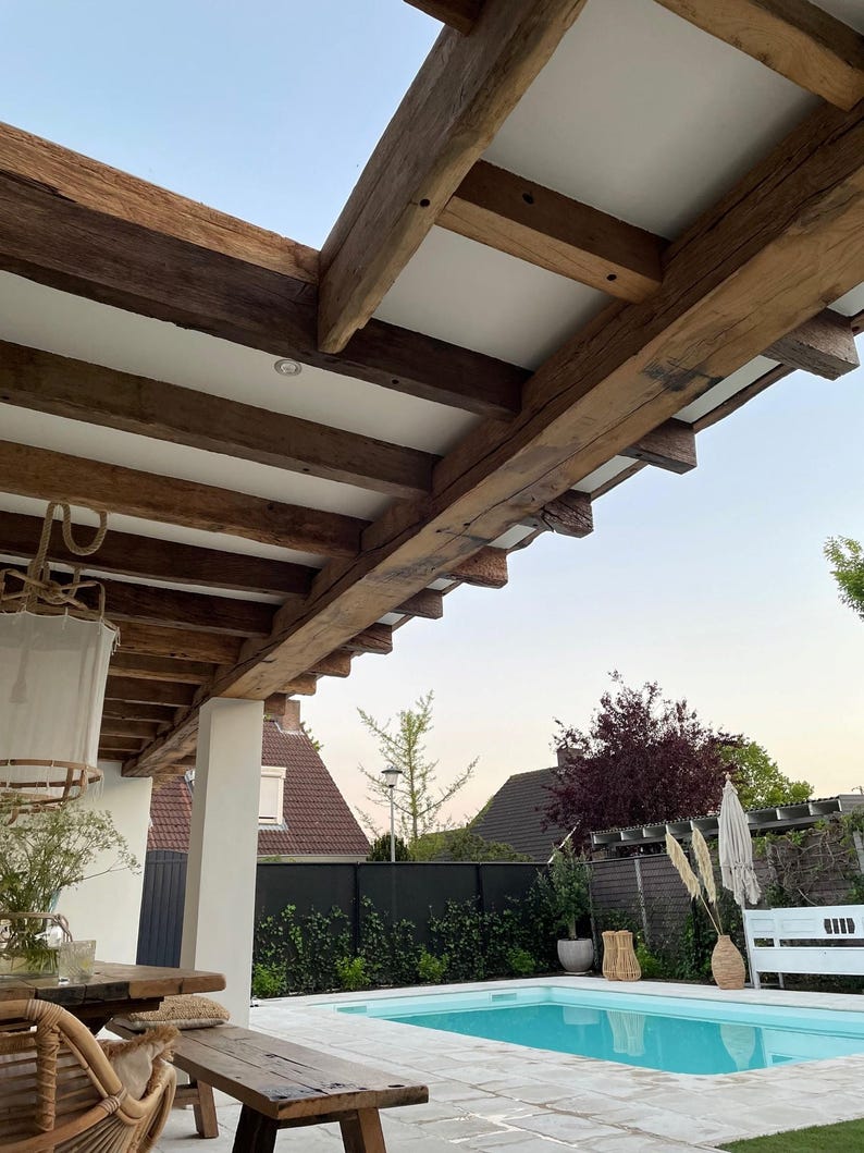 May include: An outdoor patio with a swimming pool. The patio features a wooden pergola with exposed beams and white panels. A wooden table and bench are in the foreground. The pool has clear blue water, and the background includes houses and greenery.