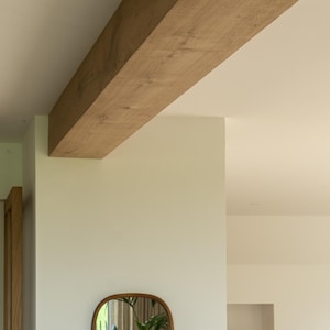 Puede incluir: Una viga de madera marr&oacute;n claro se extiende por la parte superior de la imagen, sostenida por una pared blanca. Un espejo ovalado con marco marr&oacute;n cuelga de la pared, reflejando una planta verde. La habitaci&oacute;n tiene un dise&ntilde;o minimalista.