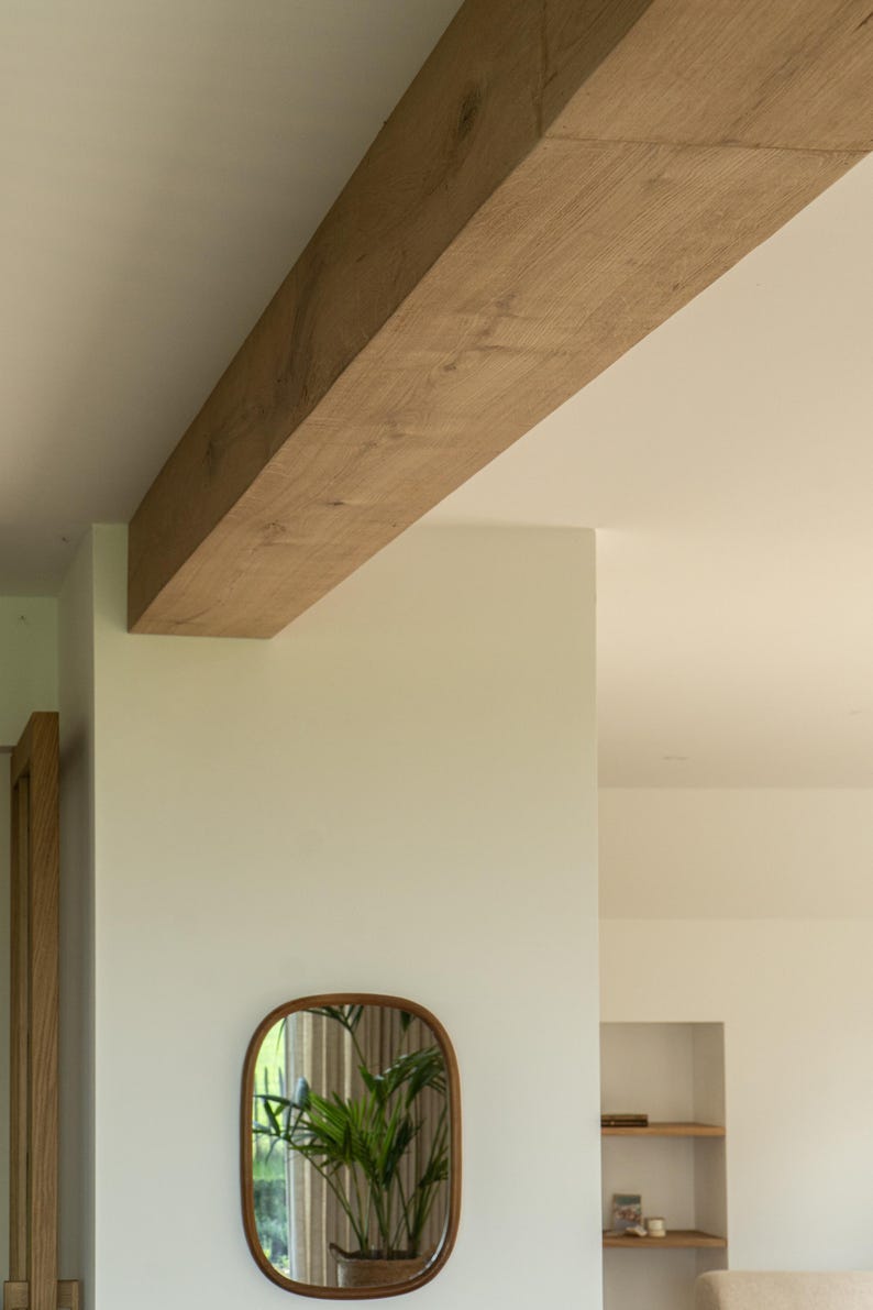 Puede incluir: Una viga de madera marr&oacute;n claro se extiende por la parte superior de la imagen, sostenida por una pared blanca. Un espejo ovalado con marco marr&oacute;n cuelga de la pared, reflejando una planta verde. La habitaci&oacute;n tiene un dise&ntilde;o minimalista.