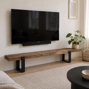 May include: A long, rectangular bench made from a thick, weathered piece of reclaimed brown wood with a rough, textured surface. The bench is supported by two U-shaped black metal legs attached to each end of the wooden beam.