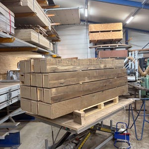 Puede incluir: Una pila de vigas de madera rectangulares y desgastadas sobre un pal&eacute;, probablemente para la construcci&oacute;n. Las vigas son de color marr&oacute;n natural, mostrando la veta de la madera. Est&aacute;n apiladas sobre una mesa elevadora en un taller.