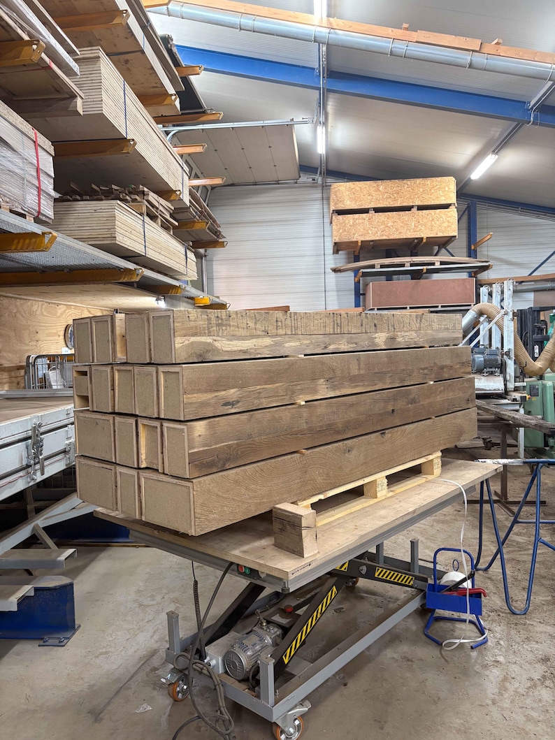 Puede incluir: Una pila de vigas de madera rectangulares y desgastadas sobre un pal&eacute;, probablemente para la construcci&oacute;n. Las vigas son de color marr&oacute;n natural, mostrando la veta de la madera. Est&aacute;n apiladas sobre una mesa elevadora en un taller.