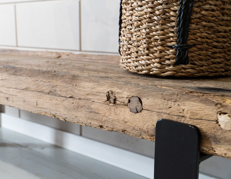 Puede incluir: Un estante de madera r&uacute;stica con una superficie desgastada y texturizada que presenta nudos y vetas naturales. El estante est&aacute; sostenido por un soporte de metal negro liso y sostiene una cesta de pasto marino tejida con detalles de cuerda negra.