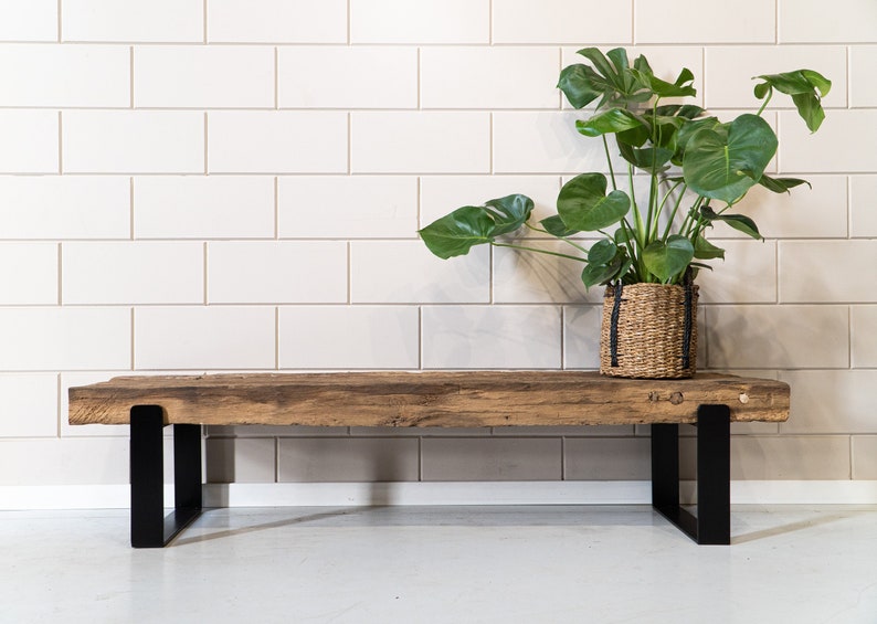 Puede incluir: Un banco rectangular con un asiento grueso de madera recuperada en tonos naturales con una superficie rugosa y texturizada. El asiento est&aacute; sostenido por dos patas de metal negro mate en forma de U fijadas en cada extremo.