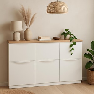 Op de afbeelding: Een witte kast met zes lades en een licht houten blad. Een beige lamp, vaas met gedroogde planten, boeken en een kleine kom staan erop. Een geweven hanglamp hangt erboven en een potplant staat rechts.