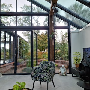 Puede incluir: Un sol&aacute;rium moderno con grandes ventanas y puertas de vidrio, que ofrece vistas a un exuberante jard&iacute;n. Una butaca estampada se encuentra en primer plano. La habitaci&oacute;n cuenta con una pared de ladrillo, vigas de madera y una chimenea negra.
