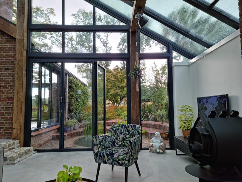 Puede incluir: Un sol&aacute;rium moderno con grandes ventanas y puertas de vidrio, que ofrece vistas a un exuberante jard&iacute;n. Una butaca estampada se encuentra en primer plano. La habitaci&oacute;n cuenta con una pared de ladrillo, vigas de madera y una chimenea negra.