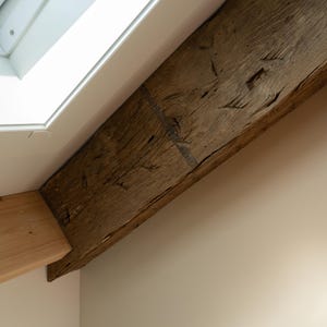 Puede incluir: Primer plano de una viga de madera marr&oacute;n oscura y desgastada, en &aacute;ngulo a trav&eacute;s del marco. La viga tiene vetas y nudos visibles, lo que sugiere edad y textura natural. Un techo blanco y una pared de color claro est&aacute;n en el fondo.