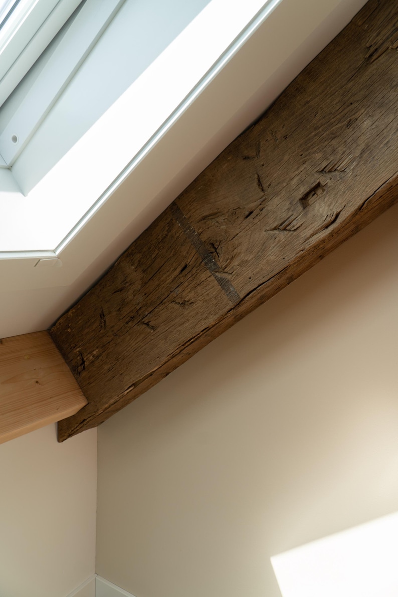 Puede incluir: Primer plano de una viga de madera marr&oacute;n oscura y desgastada, en &aacute;ngulo a trav&eacute;s del marco. La viga tiene vetas y nudos visibles, lo que sugiere edad y textura natural. Un techo blanco y una pared de color claro est&aacute;n en el fondo.