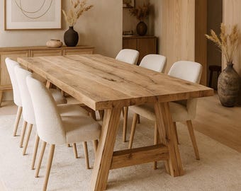 May include: A large, rectangular wooden dining table with a natural finish, supported by sturdy, angled legs. Six cream-colored upholstered chairs surround the table. The room features neutral tones, a rug, and decorative vases.