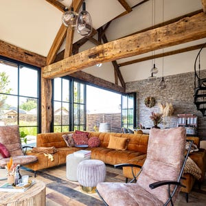 May include: A spacious living room with a high, exposed beam ceiling and large windows. The room features a brown sectional sofa, armchairs, and a wooden coffee table. Decorative pillows and pendant lights add to the cozy atmosphere.