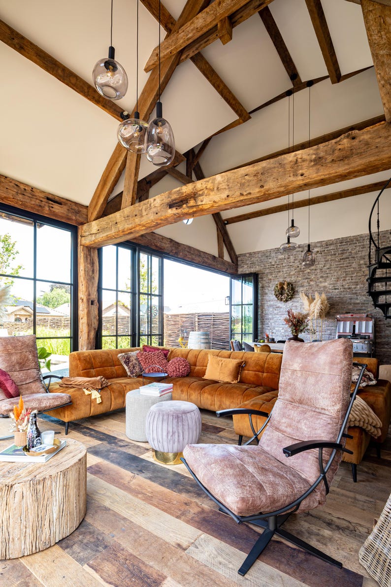 May include: A spacious living room with a high, exposed beam ceiling and large windows. The room features a brown sectional sofa, armchairs, and a wooden coffee table. Decorative pillows and pendant lights add to the cozy atmosphere.
