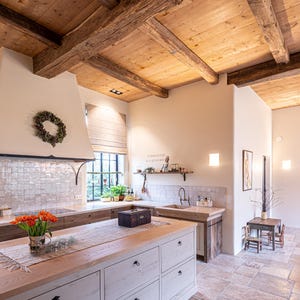 May include: A rustic kitchen with a light wood ceiling and exposed beams. The kitchen features a large island with white cabinets and a wooden countertop. A bouquet of orange tulips sits on the island. The floor is made of stone tiles.