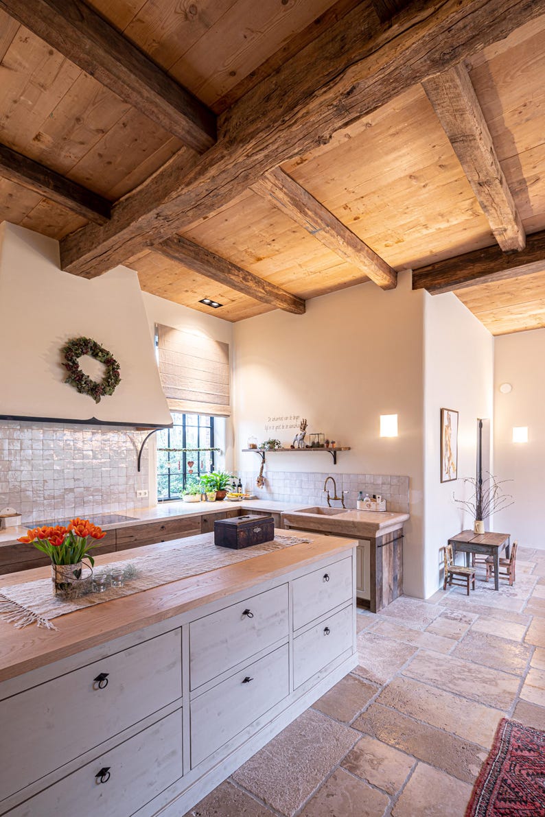 May include: A rustic kitchen with a light wood ceiling and exposed beams. The kitchen features a large island with white cabinets and a wooden countertop. A bouquet of orange tulips sits on the island. The floor is made of stone tiles.