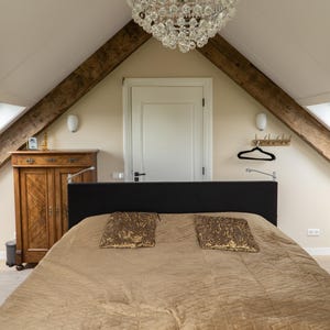 Puede incluir: Interior de dormitorio con cama, almohadas y un armario de madera. La habitaci&oacute;n tiene un techo inclinado con vigas de madera a la vista y una l&aacute;mpara de ara&ntilde;a. La cama tiene un edred&oacute;n marr&oacute;n y dos almohadas decorativas.