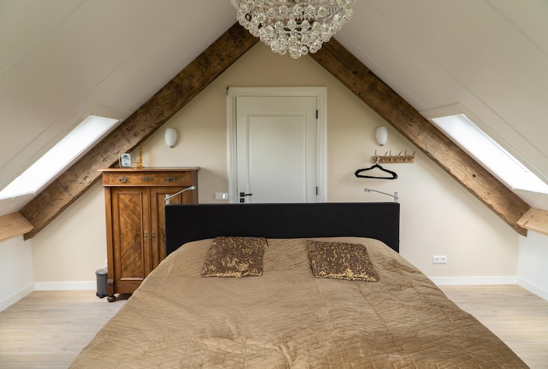 Puede incluir: Interior de dormitorio con cama, almohadas y un armario de madera. La habitaci&oacute;n tiene un techo inclinado con vigas de madera a la vista y una l&aacute;mpara de ara&ntilde;a. La cama tiene un edred&oacute;n marr&oacute;n y dos almohadas decorativas.