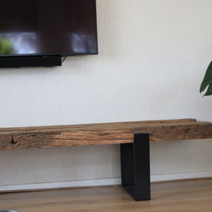 Puede incluir: Un banco largo y rectangular que presenta una viga de madera marr&oacute;n gruesa y desgastada con una superficie rugosa y texturizada con vetas visibles. El banco est&aacute; sostenido por dos patas de metal negro mate en forma de U.