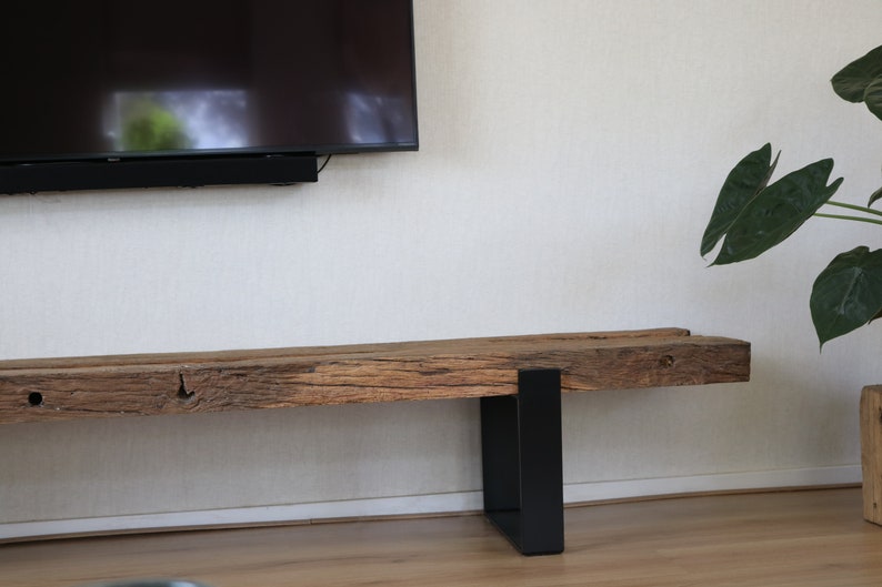 Puede incluir: Un banco largo y rectangular que presenta una viga de madera marr&oacute;n gruesa y desgastada con una superficie rugosa y texturizada con vetas visibles. El banco est&aacute; sostenido por dos patas de metal negro mate en forma de U.