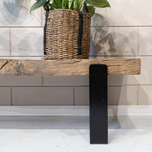 Puede incluir: Un banco de madera r&uacute;stica con un asiento de madera gruesa y desgastada con veta visible y textura natural. El banco est&aacute; sostenido por una pata de metal negro plana y vertical. Una cesta de junco marino trenzado con asas de cuerda negra descansa encima.