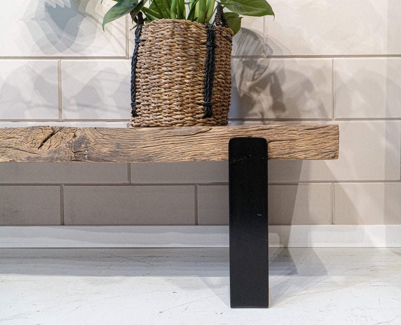 Puede incluir: Un banco de madera r&uacute;stica con un asiento de madera gruesa y desgastada con veta visible y textura natural. El banco est&aacute; sostenido por una pata de metal negro plana y vertical. Una cesta de junco marino trenzado con asas de cuerda negra descansa encima.