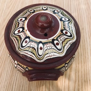 May include: A brown ceramic pot with a lid. The lid features a decorative design of white, black, and green swirls and dots. The pot has a brown handle on each side.