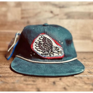 May include: Teal baseball cap with a white rope detail and a curved brim. The cap features a patch with an eagle design in black, white, and gold, outlined in red. The cap has a tag attached to the side.