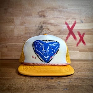 Puede incluir: Una gorra de camionero amarilla y blanca con un murciélago azul bordado en forma de corazón en la parte delantera. La gorra tiene una parte trasera de malla y un detalle de costura rosa alrededor del borde.