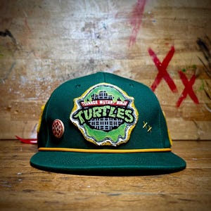 May include: A green baseball cap featuring a Teenage Mutant Ninja Turtles logo patch. The cap has a yellow rope detail and a pizza-themed button. The cap has two yellow cross-stitch details.