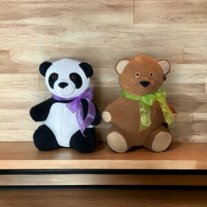 May include: Two plush stuffed animals, a black and white panda bear with a purple ribbon and a brown teddy bear with a green ribbon, sitting on a wooden shelf.
